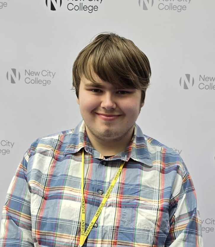 A boy wearing shirt, with yellow lanyard, looking at the camera, smiling, behind him is a build board with New City College logo