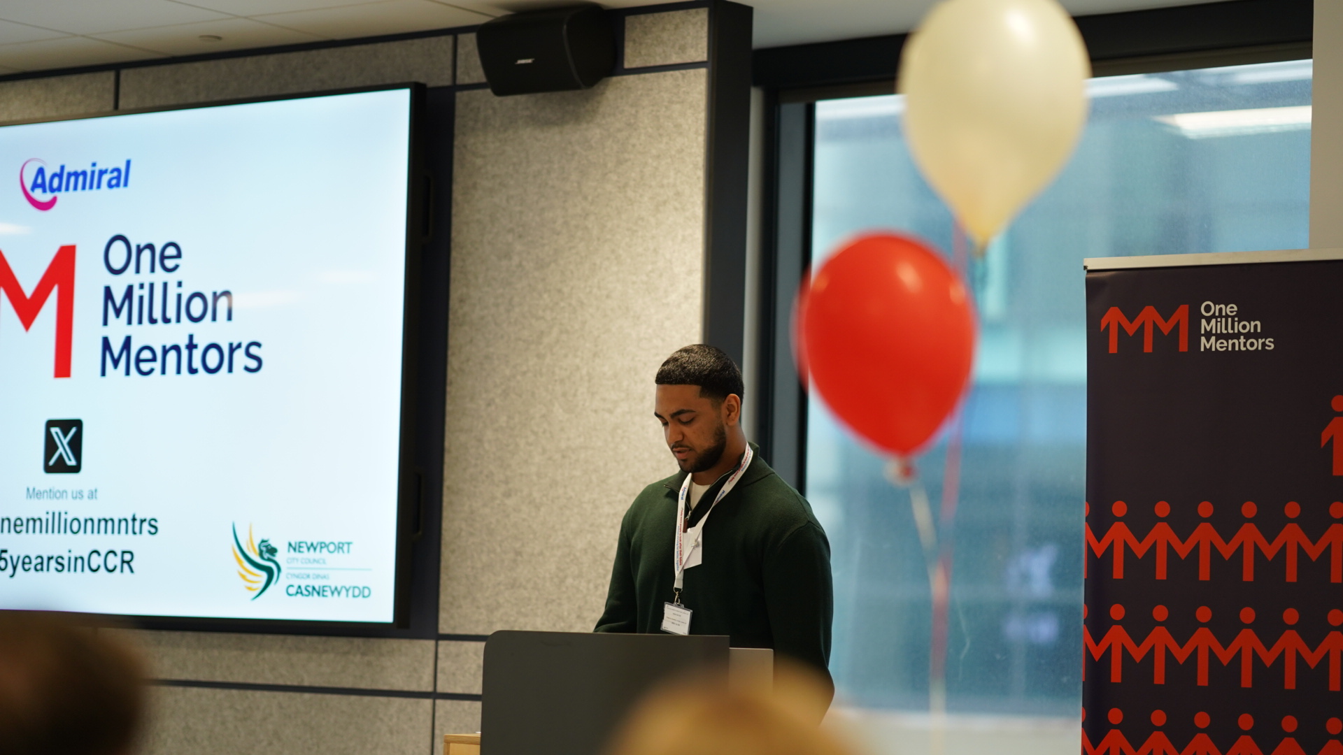 An young man, standing and giving a speech, on the right there is a 1MM banner stand, balloons in white and red